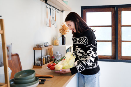 Young Asian woman stands in cozy kitchen holding bowl with fresh produce..の写真素材