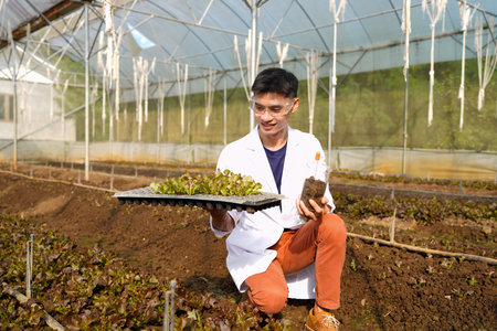 A man in a white lab coat is examining a plant and soil in a greenhouse.