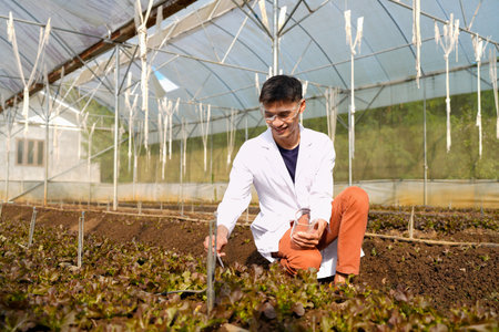 A man in a white lab coat is examining a plant and soil in a greenhouse.