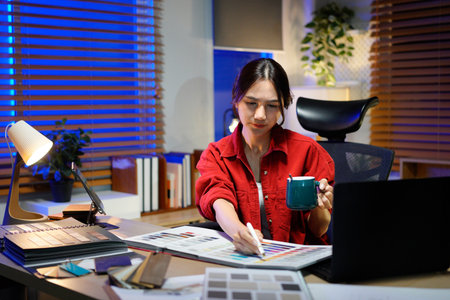 Young female architect designer working on design plans and drinking hot coffee in office at night.の写真素材