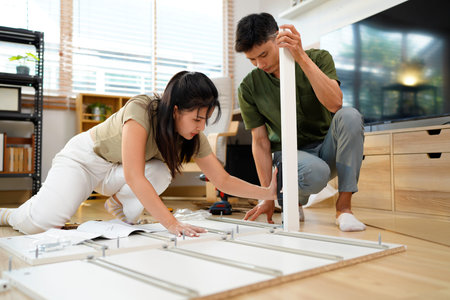 Asian adult couple assembling wooden furniture together in modern living room create new home decor...