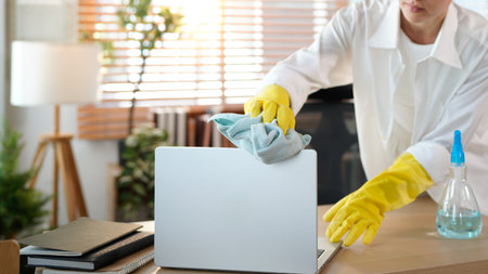 young woman housewife in yellow rubber gloves wiping dust on laptop standing in living room near...