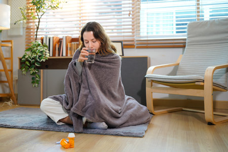 A young woman sitting on the floor with a blanket because she is sick and has a cough from the influenza while taking medicine to relieve symptoms in the living room of her home.の写真素材