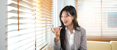 Asian businesswoman in a gray suit talking to voice assistant or chatbot on her smartphone in...