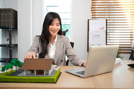 House Deal with Real Estate, An attractive, poised female agent delves into the nuances of a deal, carefully reviewing paperwork. A model house sits nearby, a tangible symbol of the subject.の写真素材