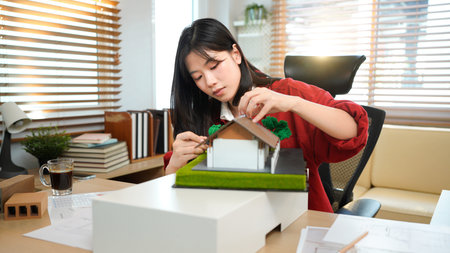 A female architect holding and working on an architecture house model with shop drawing paper in the office, Young woman architect building model house working in modern office.の写真素材