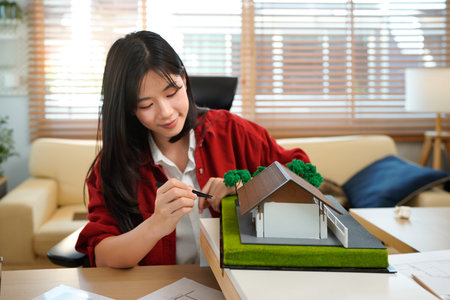 A female architect holding and working on an architecture house model with shop drawing paper in the office, Young woman architect building model house working in modern office.の写真素材
