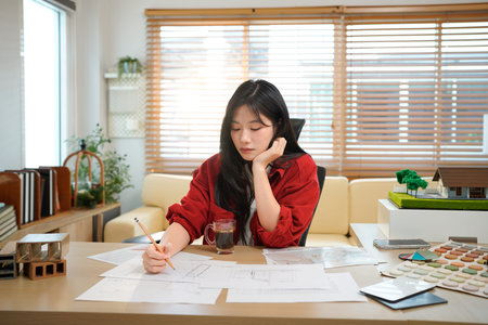 Young asian woman architect working in office with blueprints.Engineer inspect architectural plan, sketching a construction project. Portrait of woman sitting at workplace. Business concept.の写真素材