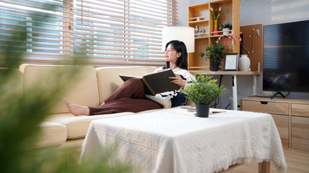 Young asian smiling cheerful woman in a warm sweater reading a book while sitting on the couch in living room at home.の写真素材