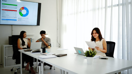 Focused female manager reading e-mails over laptop while colleagues working in the background in...