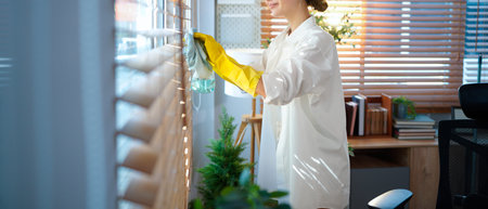 A woman cleans window blinds at home with a rag and gloves, focusing on dust removal, hygiene,...