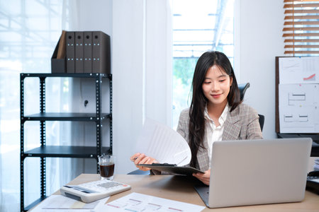 Beautiful young Asian businesswoman using laptop computer and paperworks with planning working on...