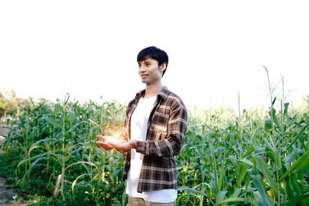 A young farmer stands in a Cornfield, wearing a plaid shirt over a white T-shirt. He smiles gently...