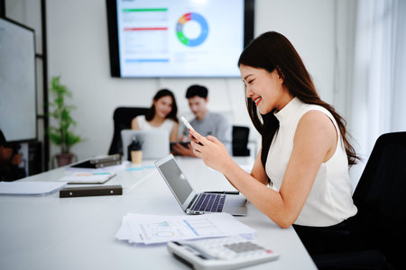 Focused businesswoman using smartphone and working over laptop in board room with colleagues in the...