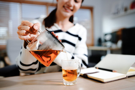 woman work study from home take break in job pouring hot tea from transparent kettle into cup,...