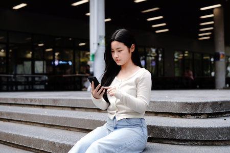 Asian student relaxing on campus stairs using smartphone.