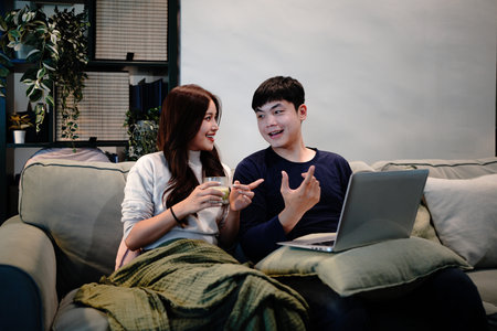 Happy young couple using laptop relaxing on couch at home. Smiling asian man and woman looking at...