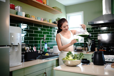 A woman in white workout attire prepares a green vegetable and fruit smoothie, pouring it into a...