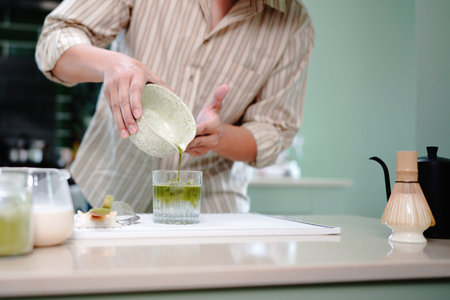 young asian man preparing iced matcha latte pouring green matcha tea into a glass with milk, creating a refreshing iced matcha latte in a cozy kitchen setting.の写真素材