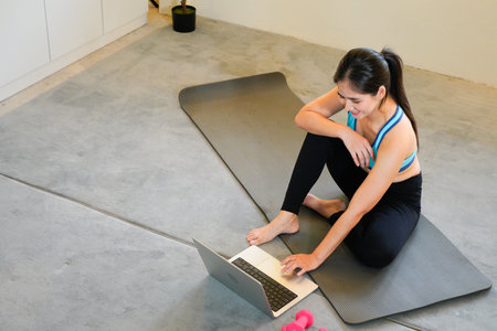 A sporty woman in sportswear is sitting on the floor with dumbbells and is using a laptop at home in the living room. Sport and recreation concept.