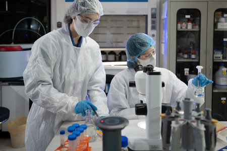Two lab scientists in full PPE collaborate over a chemical flask and microscope in Modern Research Laboratory, science and healthcare content