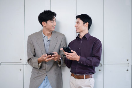 Two Asian male colleagues smiling and discussing something while looking at a cellphone, sharing information or ideas during a work break technology, communication, and business lifestyle concepts