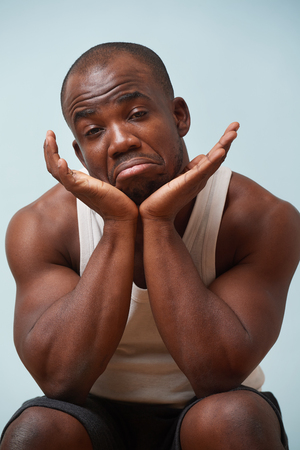 Handsome bold black man in white sleeveless shirt grimacing against pale blue background. Sitting crouched with upset sad face. Leaning chin on palms.の写真素材