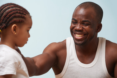 Handsome black young father cheering up his tired cute little daughter against pale blue background. Smiling for her, putting hand on her shoulderの写真素材