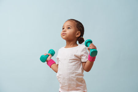 Portrait of cute little mulatto girl lifting small colored dumbbells against pale blue background. Pink shorts and wristbands. Fitness workout.の写真素材