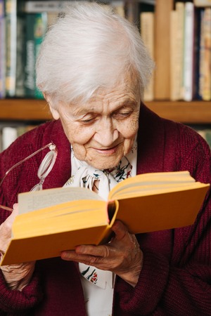 Portrait of cheerful elderly grey haired woman. She's reading, standing with book in one hand and glasses in another, bent closely to see letters better. Near bookshelf at home.の写真素材