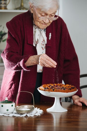 Senior gray haired woman pouring powdered sugar on tasty holiday pie on a dinner table at homeの写真素材