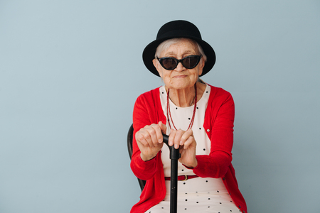Respectable well dressed in black mini hat and sunglasses gray haired fancy senior woman with cane is resting on a chair at home on a sunny spring day.の写真素材