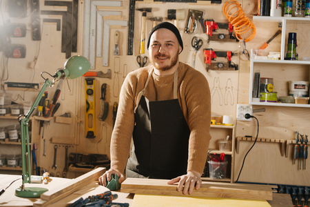 Portrait of smiling handsome middle aged bearded carpenter in a watch cap. He is making furniture on order in a workshop. Taking few seconds between things.の写真素材