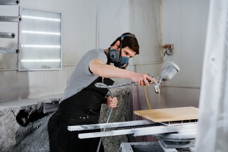 Portrait of working handsome slim middle aged brunet carpenter in respirator. He is making furniture on order in a workshop. Varnishing wood panel with HVLP spray gun.の写真素材