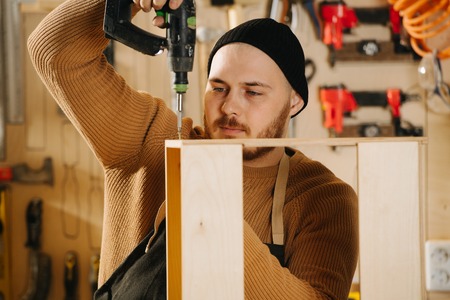 Portrait of working handsome middle aged bearded carpenter in a watch cap. He is making furniture on order in a workshop. Assemling, screwing parts of a future drawer pullout shelf.の写真素材