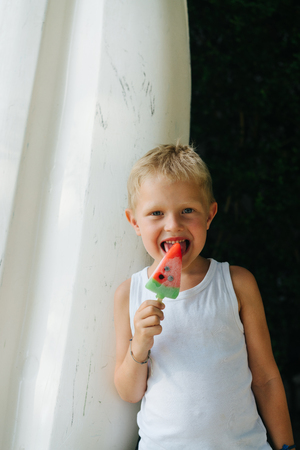 Little russian blond boy in sleeveless white shirt, licking his watermelon fruit ice pop in hot sunny summer day. Tongue out, pleased, teasing.の写真素材