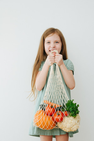 Portrait of a little girl picks up a heavy bag with vegetables on white backgroundの写真素材