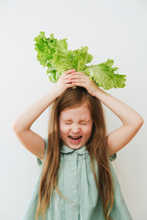 Closeup emotions portrait of girl, for fun makes the hair of green salad. At the table where a lot of vegetablesの写真素材
