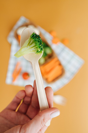 Close-up picture of broccoli on a wooden spoon in hand against the background of a plate with slice vegetablesの写真素材