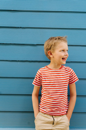 Cute little blond boy in striped T-shirt posing in front of blue house wall. He's posing, looking...