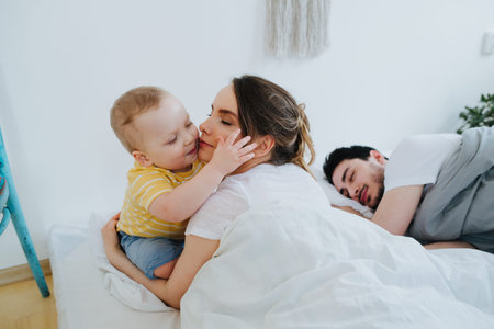 Cute little toddler son is sitting on parents bed, kissing his mother to wake her. Father still sleeping sweetly. Family morningの写真素材