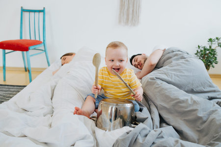 Cute little toddler son is sitting on parents bed, bashing metal pan with spoon, trying to wake his sleeping father and mother. Family morningの写真素材