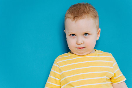 Portrait of cute shy blond little toddler boy in T-shirt looking from under eyebrows