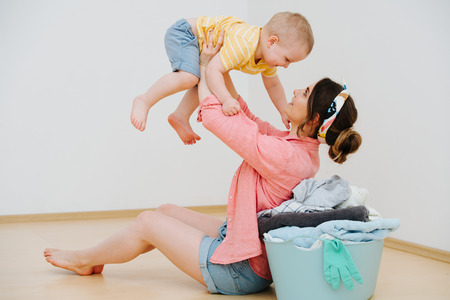 Young brunette mother sitting next to a laundry basin full of freshly washed towels. She's lifting her little son over head.の写真素材