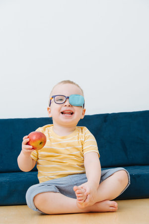 Little blond toddler boy in a lazy eye patch with an apple in hand, sitting on a floor a couch at...