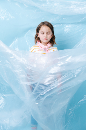 Little brunette girl is wrapped in plastic in protest of waste crisis. She closed her eyes, sinking in figurative plastic whirlpool. Recycling and ecology.の写真素材