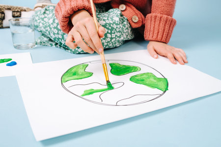 No face portrait of teenage girl painting planet earth on paper. Cropped. She uses green color on a...