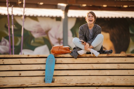 Laughing young woman with short hair in grey fashionable coat with a skateboard is sitting cross-legged on a two-stage wooden bench. lifestyle photo. Holding in her phone.の写真素材