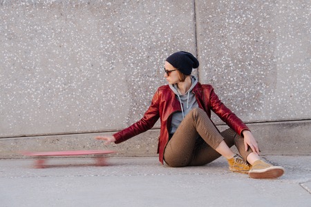 Portrait of a young happy woman with short hair in a red jacket and sunglasses. She's sitting on a street floor. Pushing her pink skateboard away.の写真素材