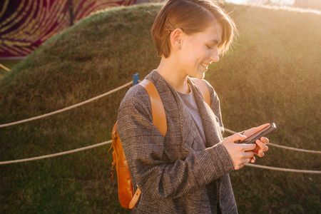 Portrait of a young happy beautiful woman with short hair in a light grey fashionable coat with a backpack in a park. She's texting in her phone.の写真素材